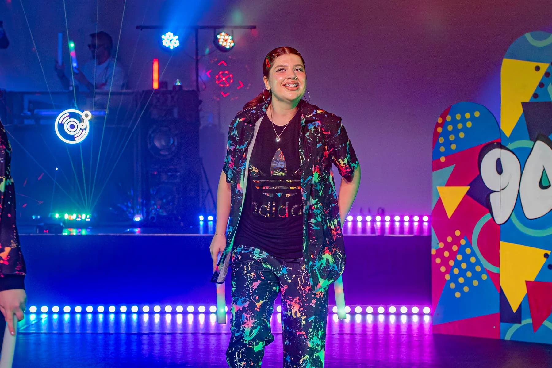 Solo performer in 90s costume on a colourful stage set.